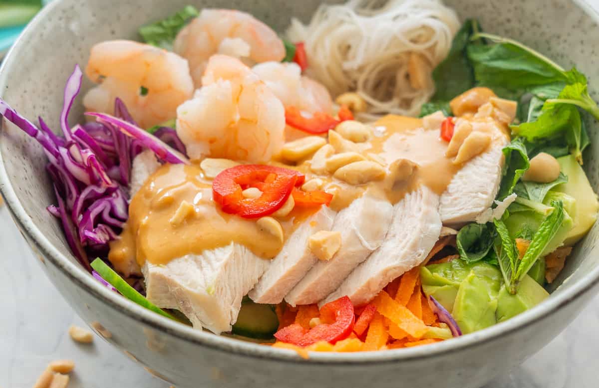 Summer roll bowl on benchtop, grated carrot, shredded cabbage, chopped lettuce, vermicelli, sliced cucumber, avocado, shrimp, chicken slices and satay sauce drizzled down the centre garnished with peanuts.