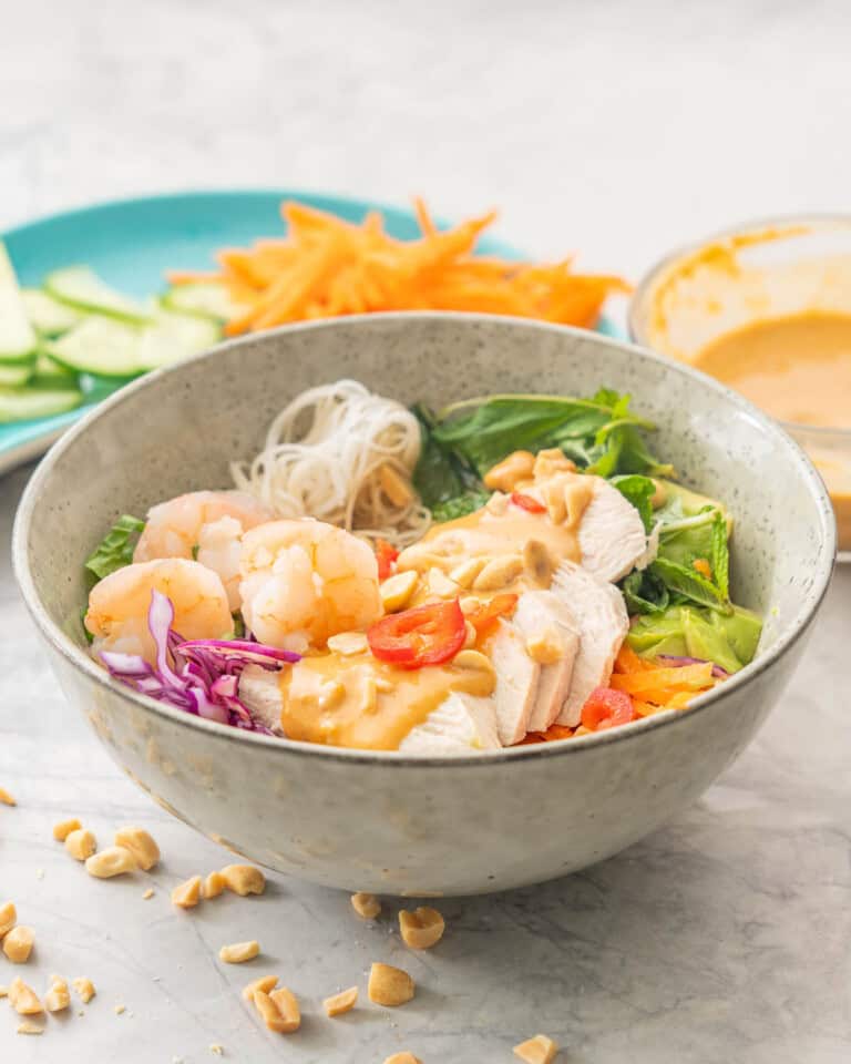 Summer roll bowl on benchtop, grated carrot, shredded cabbage, chopped lettuce, vermicelli, sliced cucumber, avocado, shrimp, chicken slices and satay sauce drizzled down the centre garnished with peanuts. Bowl in background with satay sauce and plate with grated carrot and cucmber slices.