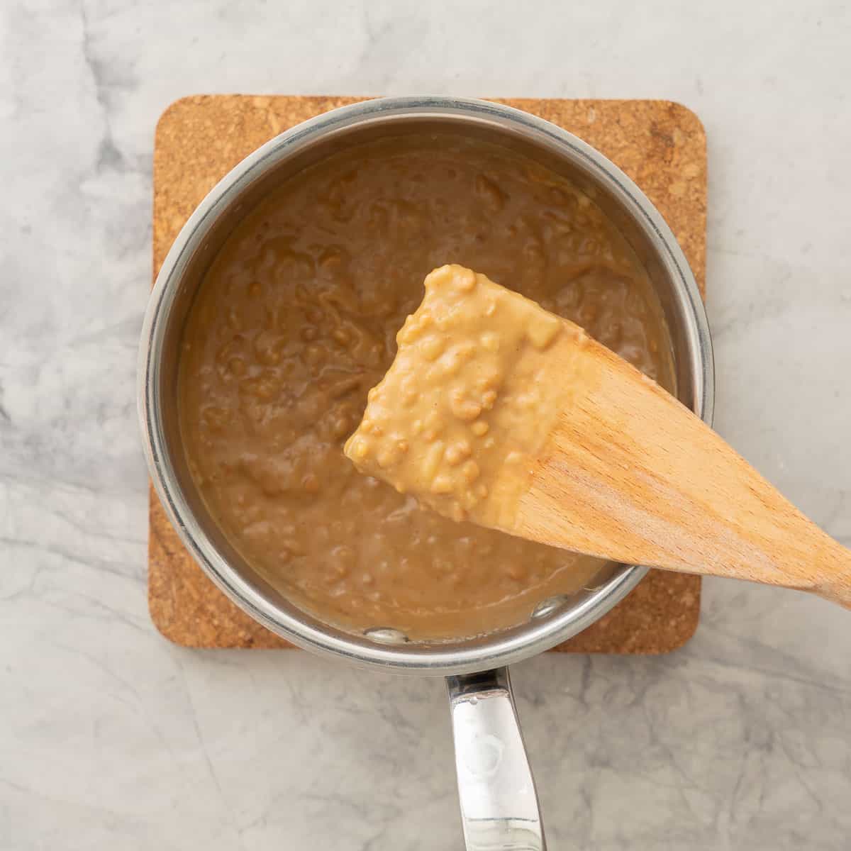 Wooden spoon scooped into Peanut Sauce held up to camera, sauce in a pot sitting on cork chopping board on bench top.