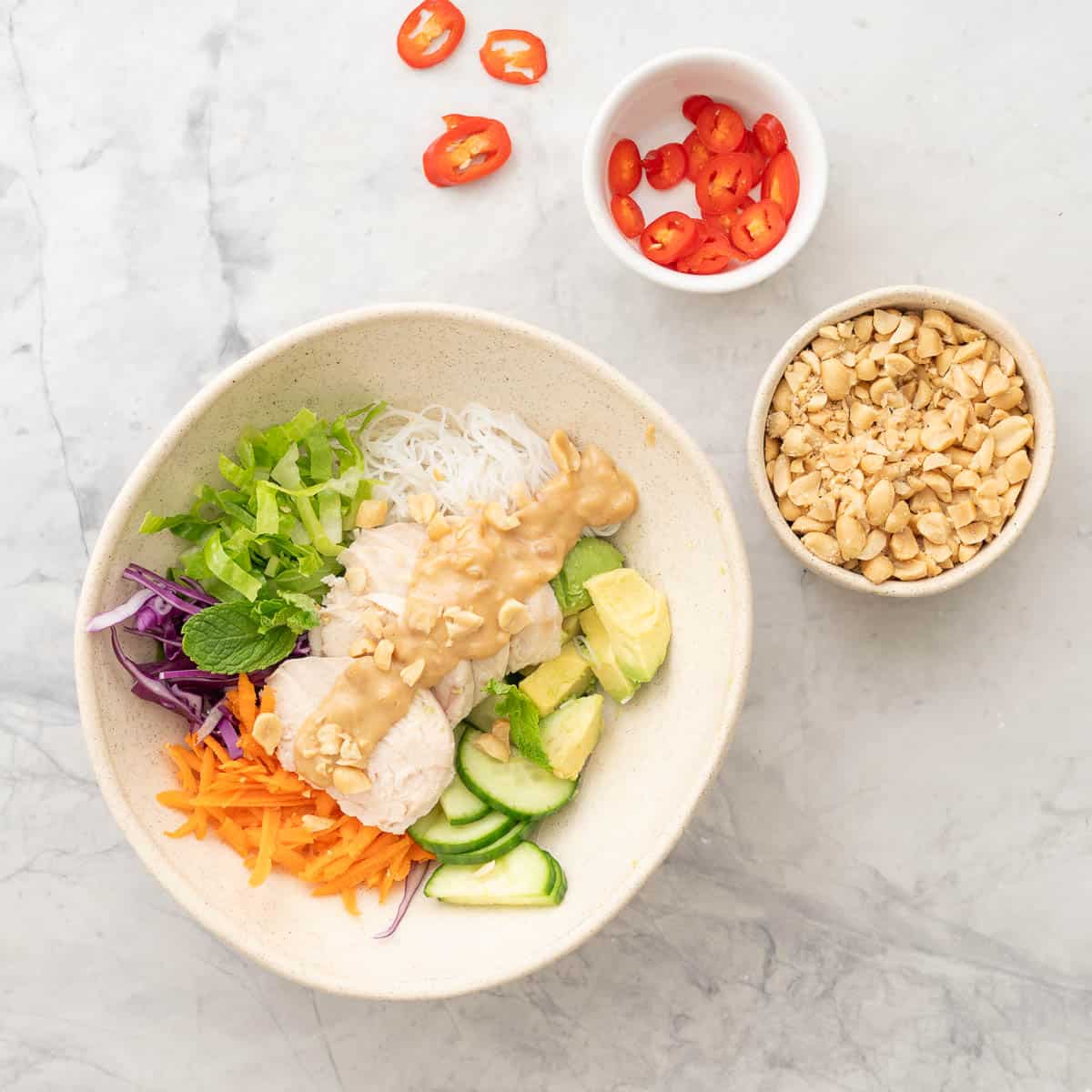 Summer roll bowl on benchtop, grated carrot, shredded cabbage, chopped lettuce, vermicelli, sliced cucumber, avocado, shrimp, chicken slices and satay sauce drizzled down the centre garnished with peanuts. Two small bowls on benchtop one with peanuts another with sliced red chilli.