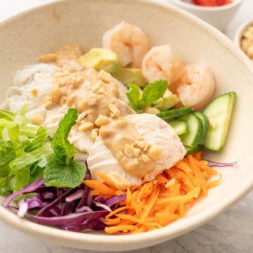 Summer roll bowl on benchtop, grated carrot, shredded cabbage, chopped lettuce, vermicelli, sliced cucumber, avocado, shrimp, chicken slices and satay sauce drizzled down the centre garnished with peanuts.