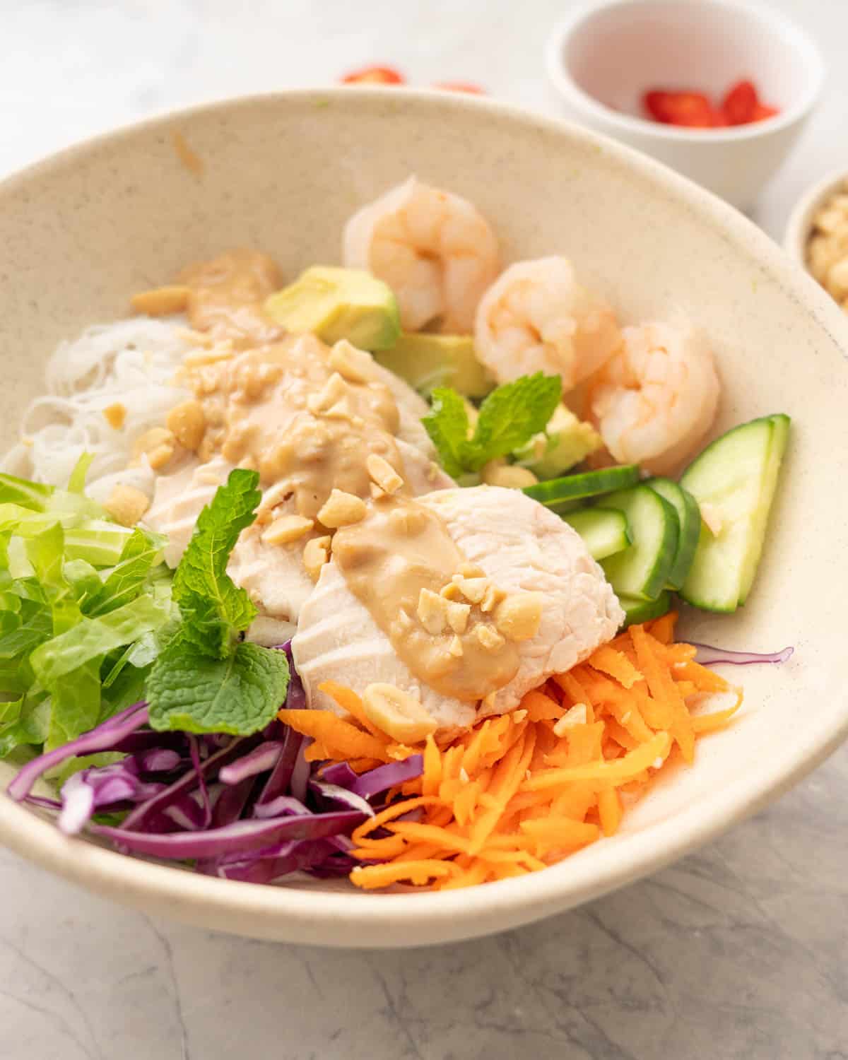 Summer roll bowl on benchtop, grated carrot, shredded cabbage, chopped lettuce, vermicelli, sliced cucumber, avocado, shrimp, chicken slices and satay sauce drizzled down the centre garnished with peanuts.