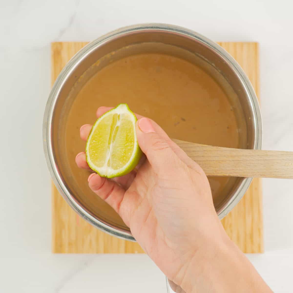 half a lime being held up above a sauce pan of smooth peanut sauce. 