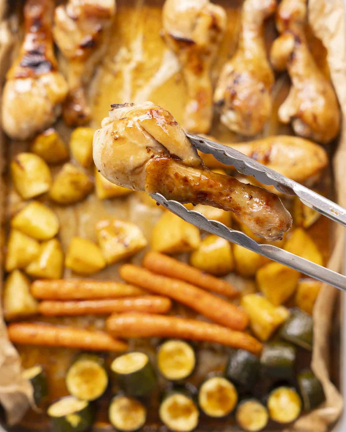 Roasted chicken glazed in honey soy sauce held up to camera with tongs, above a chicken and veggies sheet pan. 