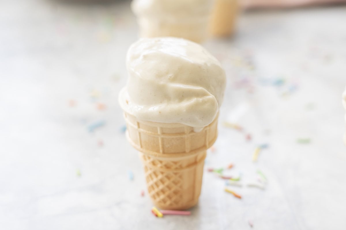 A mini ice cream cone on benchtop with Banana ice cream scooped on top, coloured sprinkles on bench.