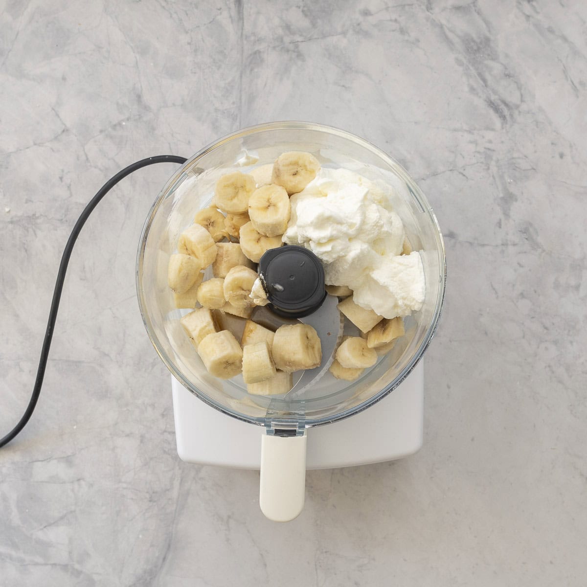 Frozen banana pieces, greek yogurt and vanilla essence in a food processor on bench top.