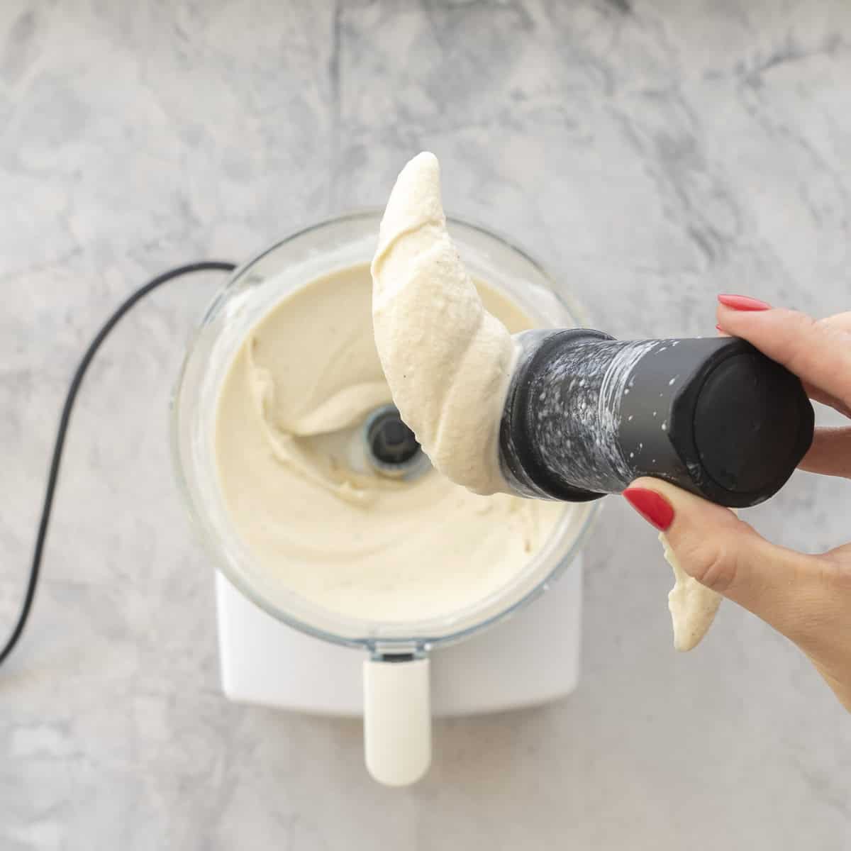 Hand holding food processor blade with thick creamy banana ice cream on it food processor with banana ice cream below on bench.