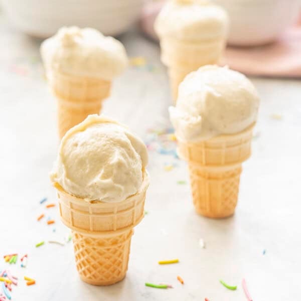 Four mini ice cream cones on benchtop with Banana ice cream scooped on top, coloured sprinkles on bench.