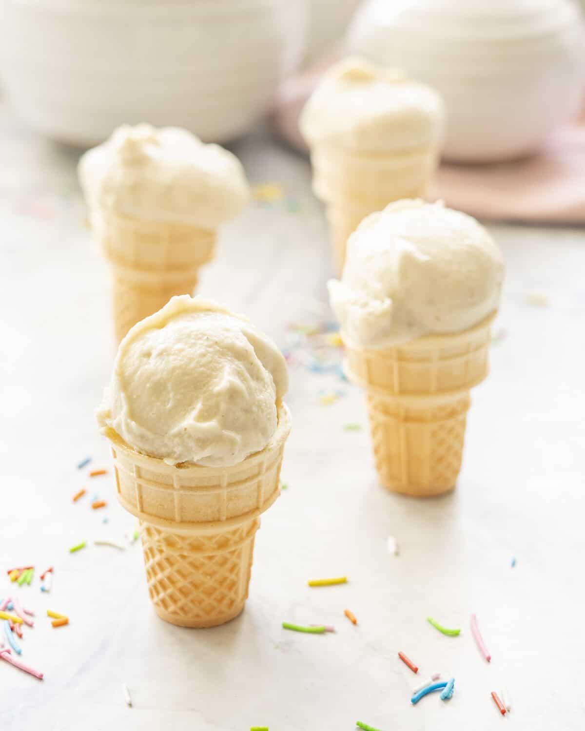 Four mini ice cream cones on benchtop with Banana ice cream scooped on top, coloured sprinkles on bench.