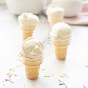 Four mini ice cream cones on benchtop with Banana ice cream scooped on top, coloured sprinkles on bench.