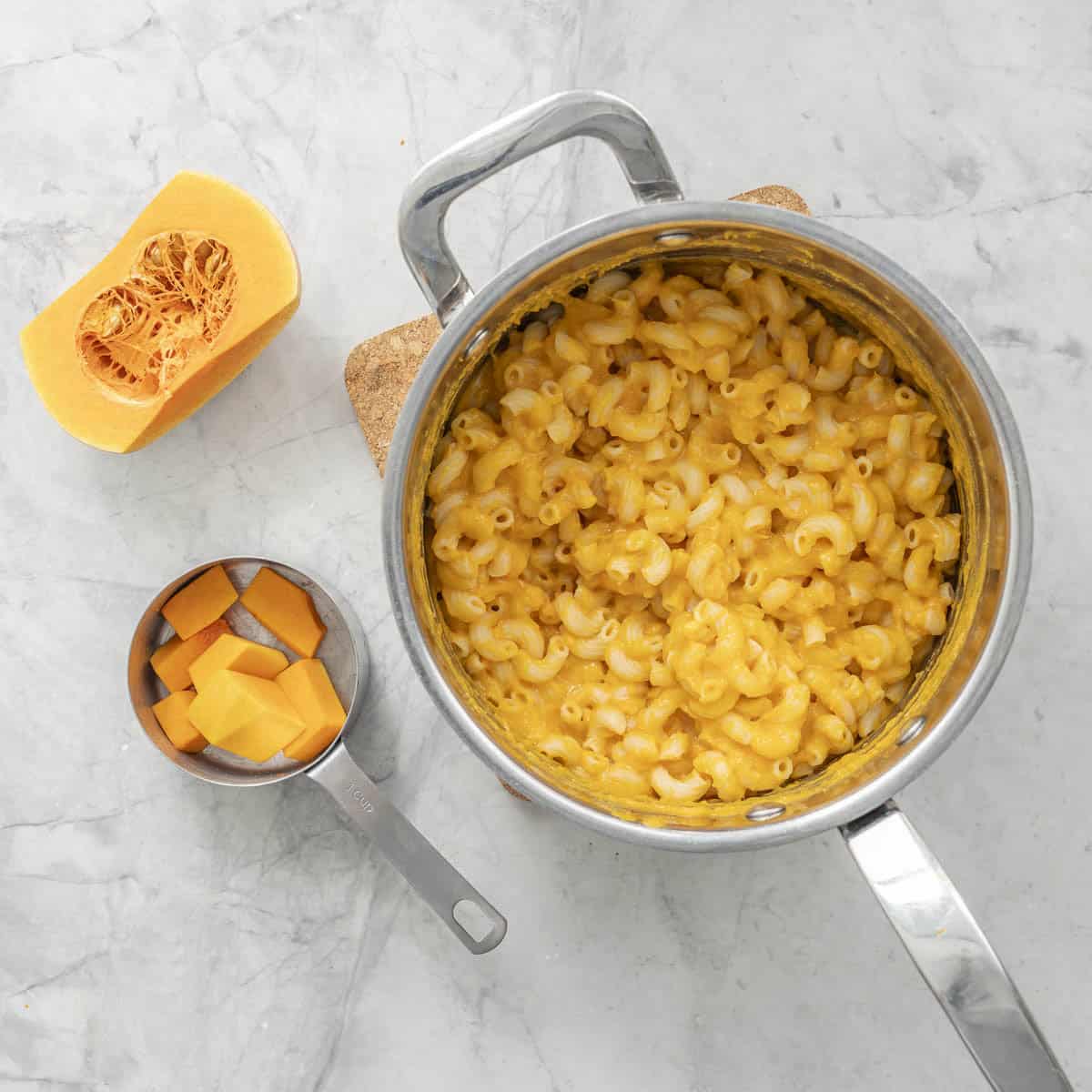 Butternut macaroni and cheese in sauce pan on bench top, half a butternut on benchtop, measuring cup with chopped up butternut pieces inside.