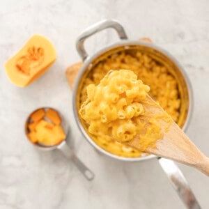 A scoop of butternut macaroni and cheese on wooden spoon held up above sauce pan of butternut macaroni and cheese, half a butternut and measuring cup with chopped butternut inside.