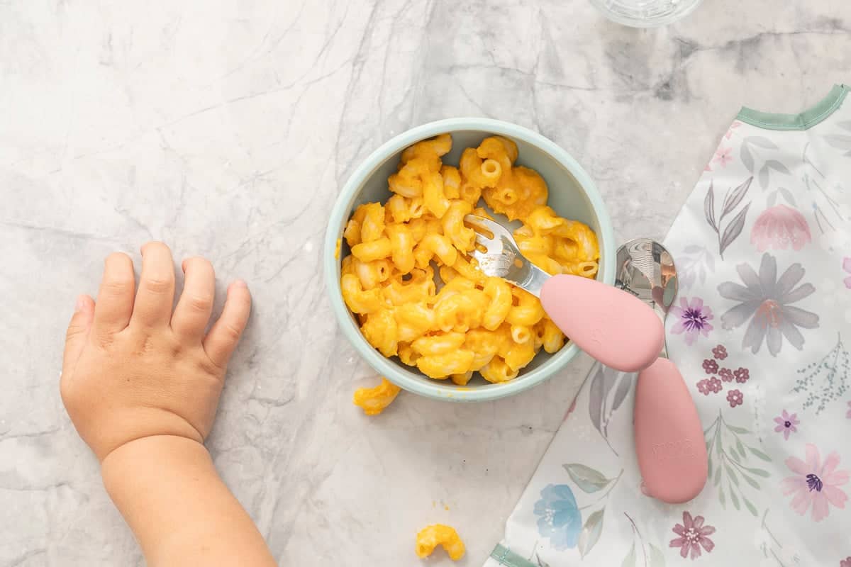 Silicone baby bowl with butternut mac and cheese inside, pink handled silicone toddler fork inside bowl, toddlers hand resting on benchtop.