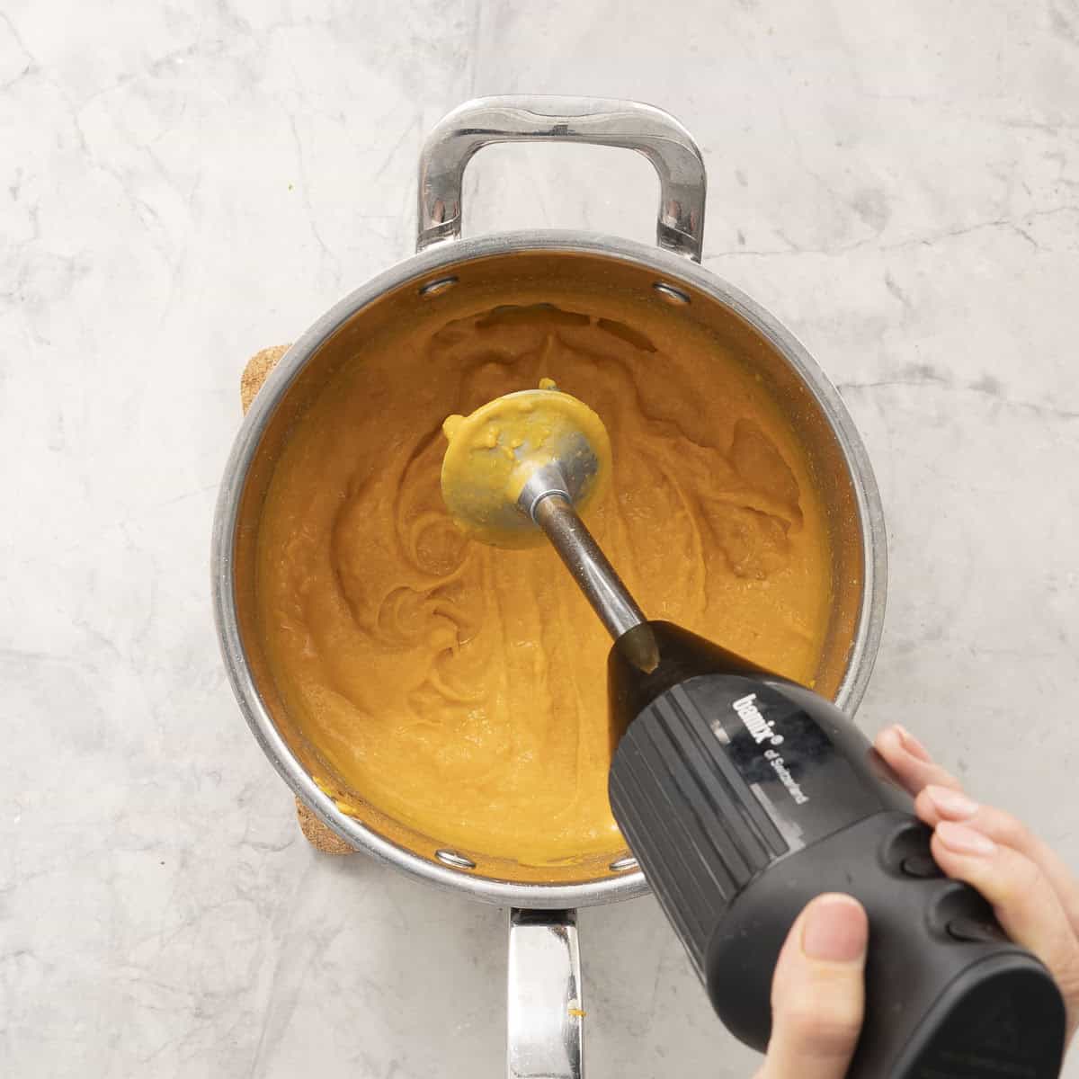 Hand blender blending together butternut and milk until smooth in sauce pan on benchtop.