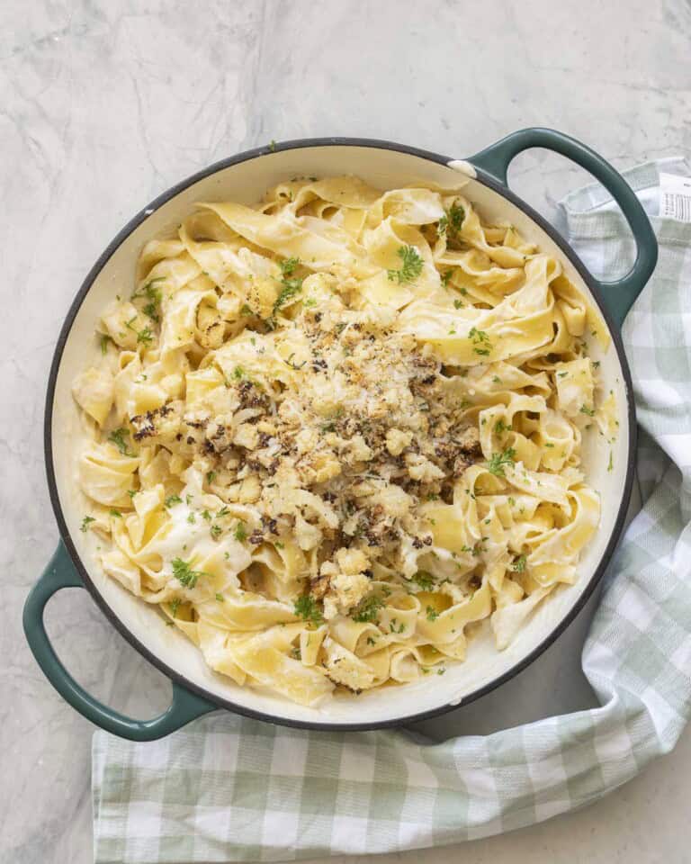 Cauliflower alfredo sauce mixed through cooked pasta topped with crispy roasted cauliflower, panko bread crumbs, thyme, parsley and parmesan cheese in a ceramic dutch oven pan on benchtop with green and white checkered tea towel.