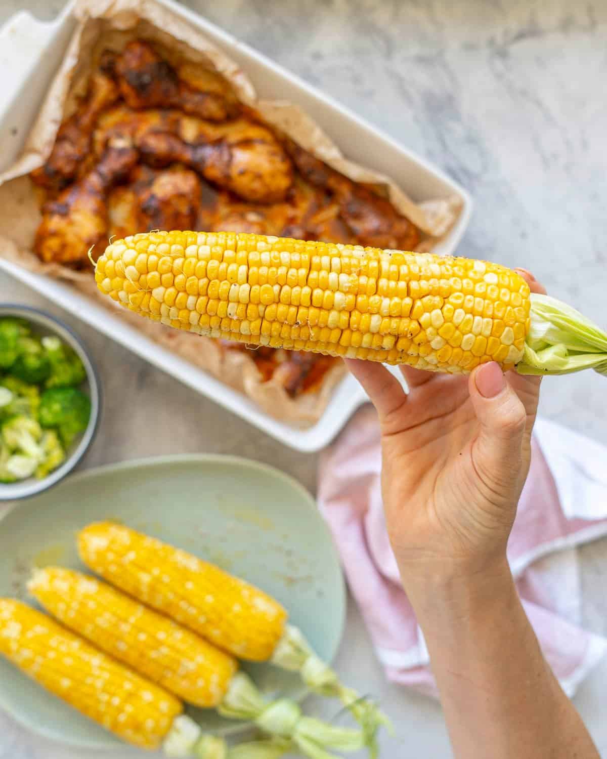 Hand holding a cooked corn on the cob with melted butter, salt and pepper on top above a white baking dish with cooked chicken drumsticks inside, bowl of cooked broccoli florets and plate with three corn on the cob.