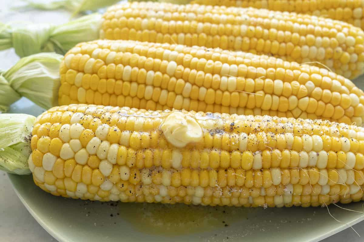 Four cooked corn on the cob on a plate with melted butter and salt and pepper on top.