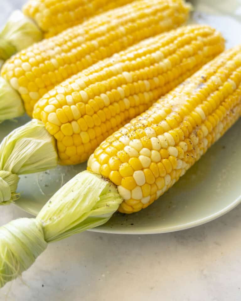 Four cooked corn on the cob on a plate with melted butter and salt and pepper on top.