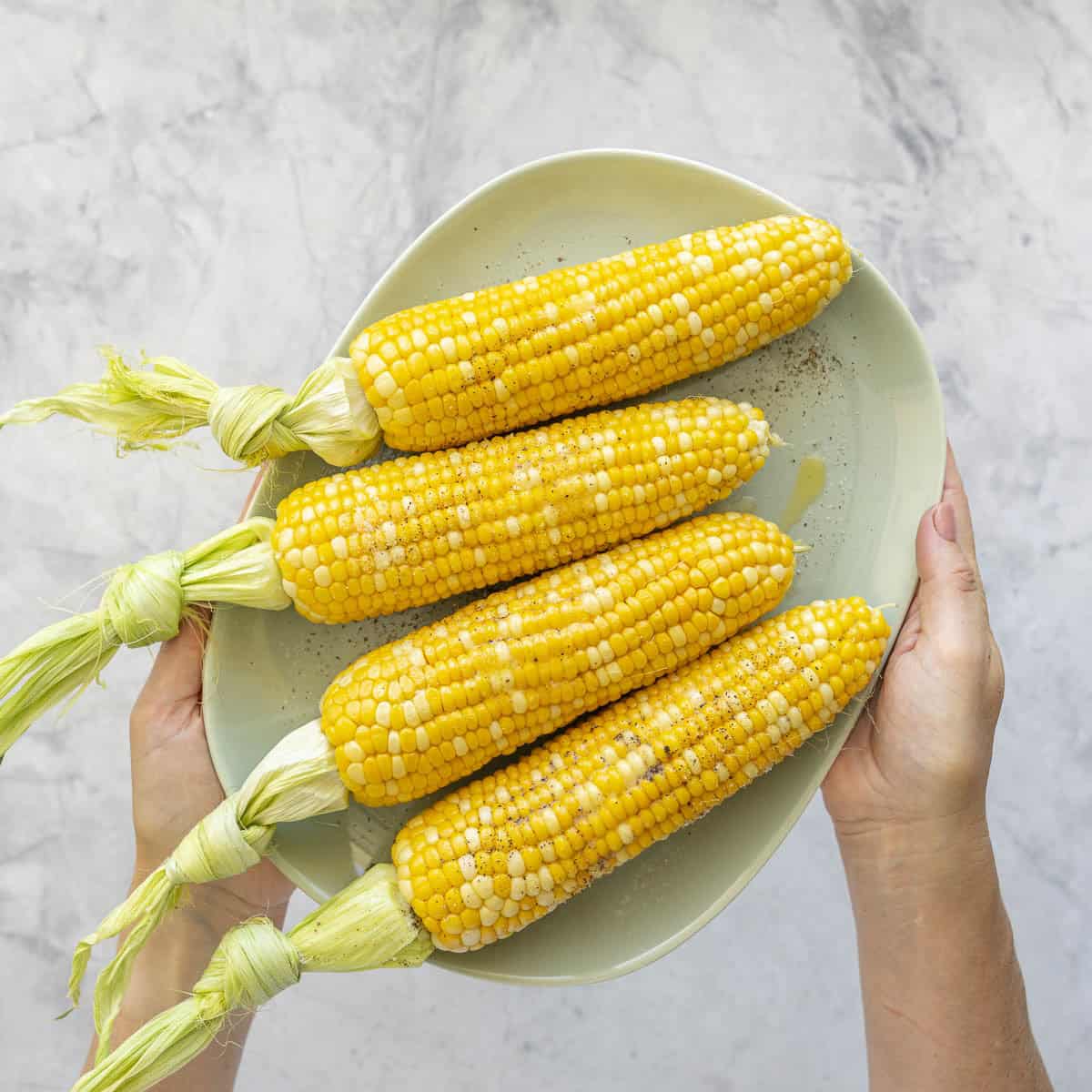 Hands holding up a plate with four cooked corn on the cob, melted butter and salt and pepper on top.