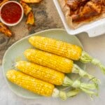 Four cooked corn on the cob on a plate with melted butter and salt and pepper on top, tray of crispy potato wedges with sweet chilli sauce, chicken drumsticks in baking tray.