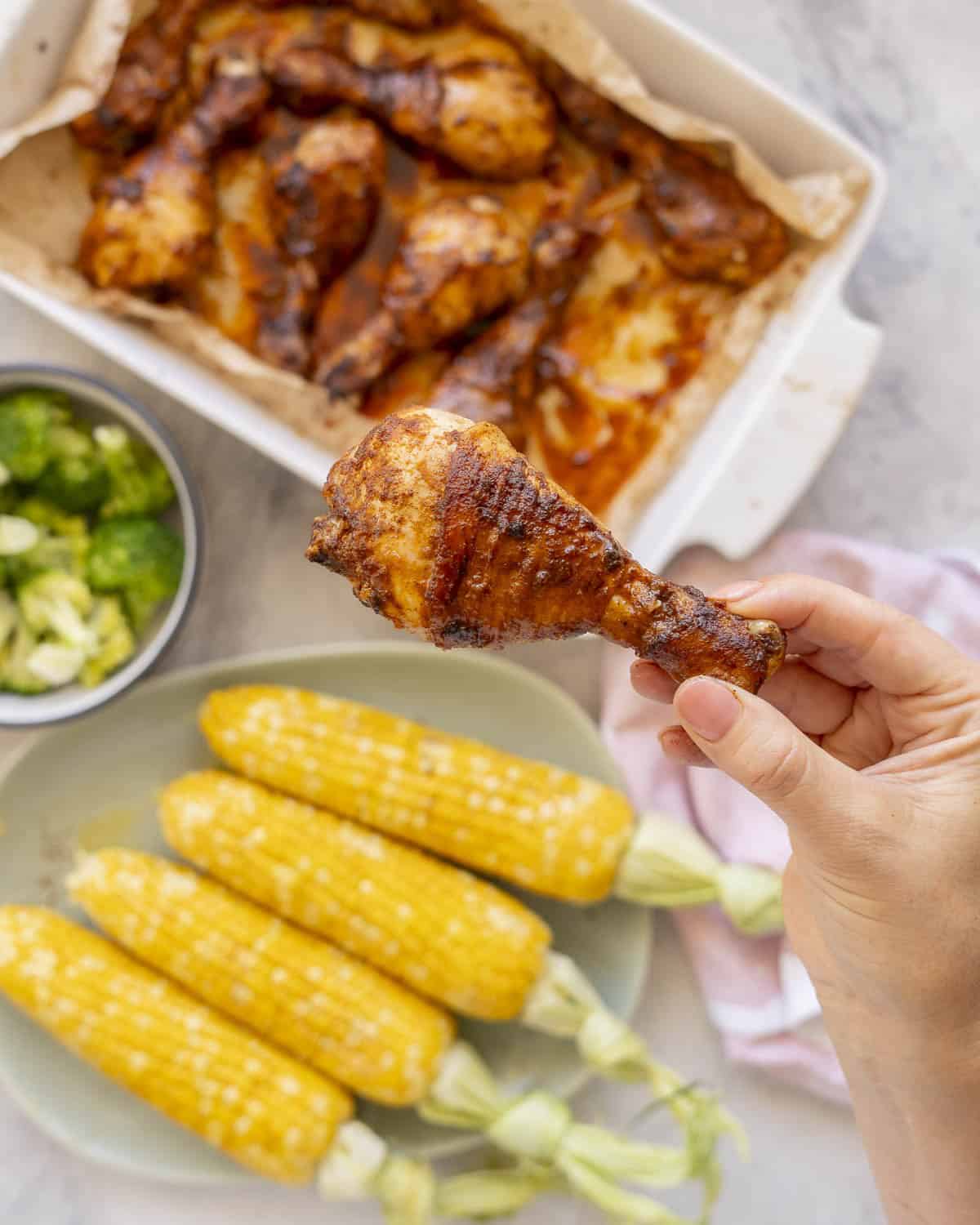 Hand holding up a glossy sticky drumstick above a white baking dish with cooked chicken drumsticks inside, bowl of cooked broccoli florets and four corn on the cob on a plate.