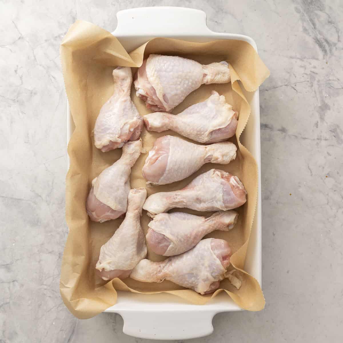 Nine chicken drumsticks inside a white oven dish lined with baking paper on benchtop.