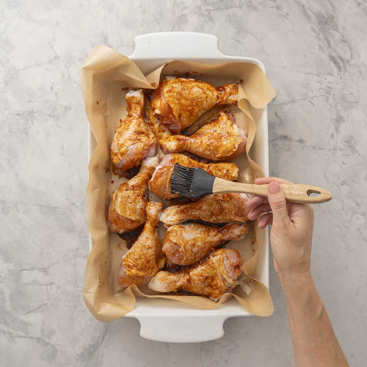 Nine chicken drumsticks inside a white oven dish lined with baking paper, hand holding a basting brush covering drumsticks with paprika mixture.