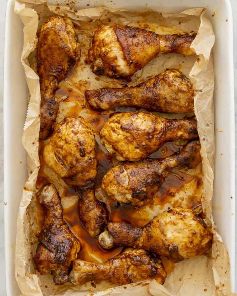 Nine cooked chicken drumsticks with sticky glossy paprika sauce on top inside a white oven dish lined with baking paper.