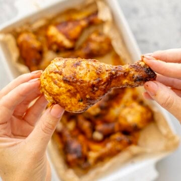 Hand holding up a glossy sticky drumstick above a white baking dish with cooked chicken drumsticks inside.