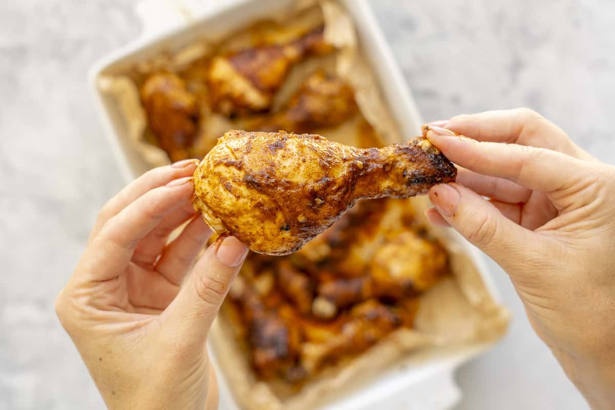 Hand holding up a glossy sticky drumstick above a white baking dish with cooked chicken drumsticks inside.