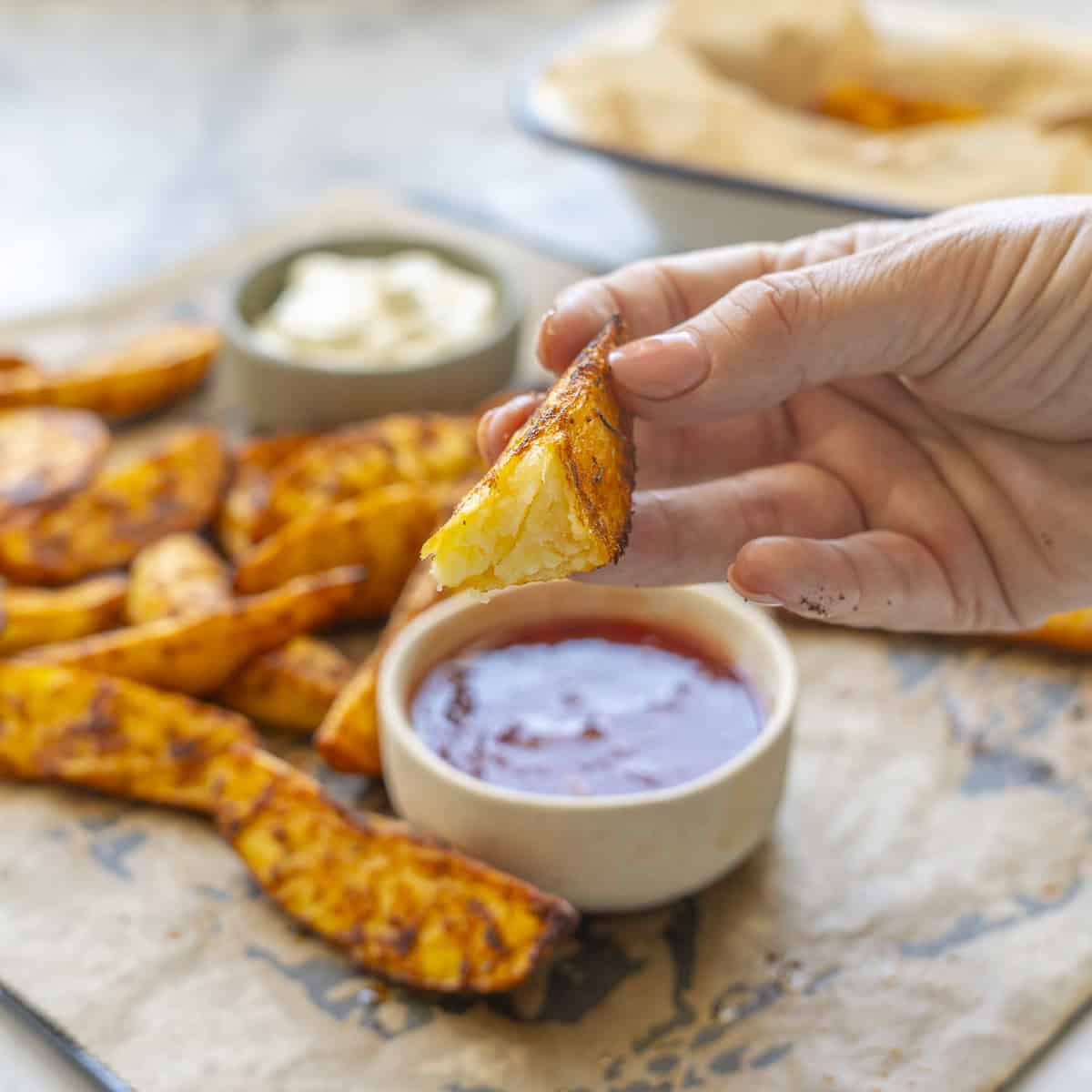 Hand holding a broken in hand potato wedge showing the inside, crispy cooked potato wedges on baking tray lined with baking paper, ramekins with aioli and sweet chilli sauce on tray.