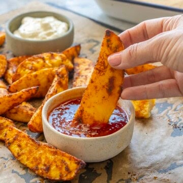 Crispy seasoned potato wedges on lined baking tray with two ramekins, aioli and sweet chilli sauce, hand holding a wedge dipping into sweet chilli sauce.