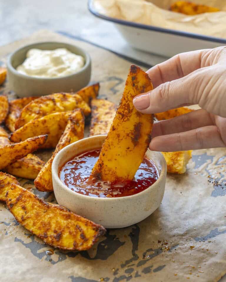 Crispy seasoned potato wedges on lined baking tray with two ramekins, aioli and sweet chilli sauce, hand holding a wedge dipping into sweet chilli sauce.