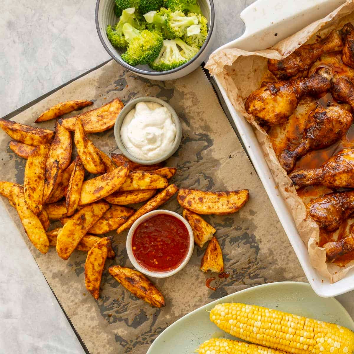 Crispy cooked potato wedges on baking tray lined with baking paper, ramekins with aioli and sweet chilli sauce on tray. White baking dish with cooked chicken drumsticks inside, bowl of cooked broccoli florets and four corn on the cob on a plate.