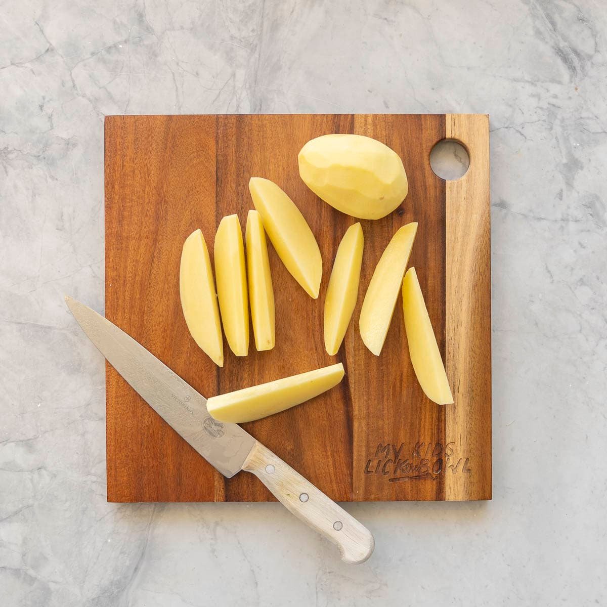 One peeled potato on wooden chopping board, potato chopped into wedge slices with knife laying on board.