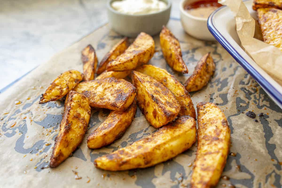 Crispy cooked potato wedges on baking tray lined with baking paper, ramekins with aioli and sweet chilli on tray.