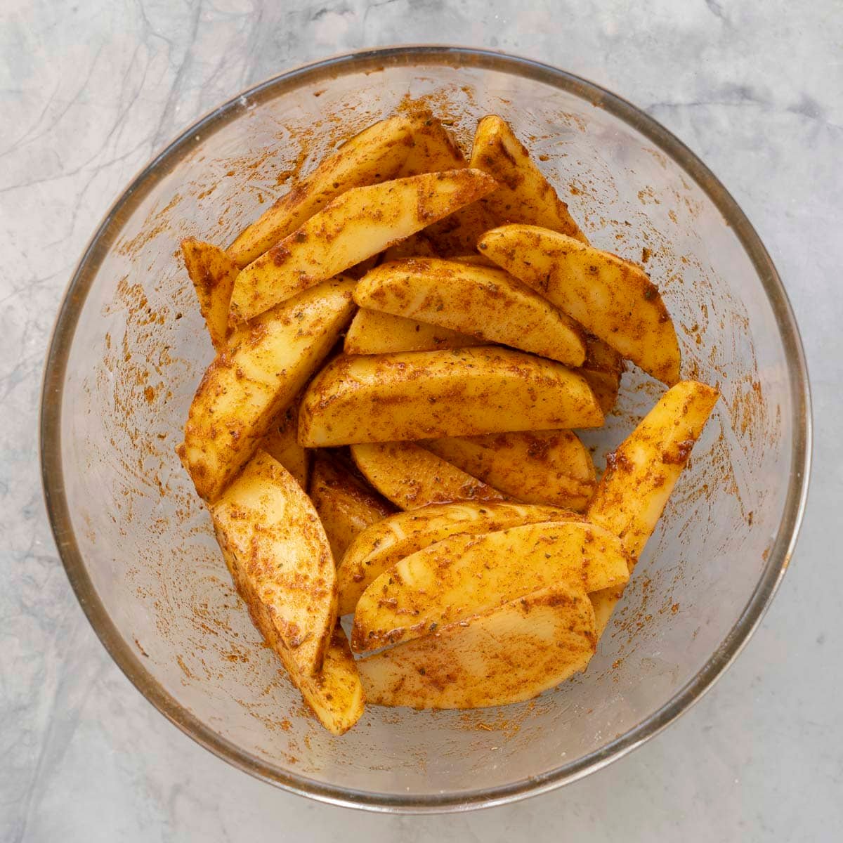 Potato wedges coated in seasonings in a glass bowl on benchtop.