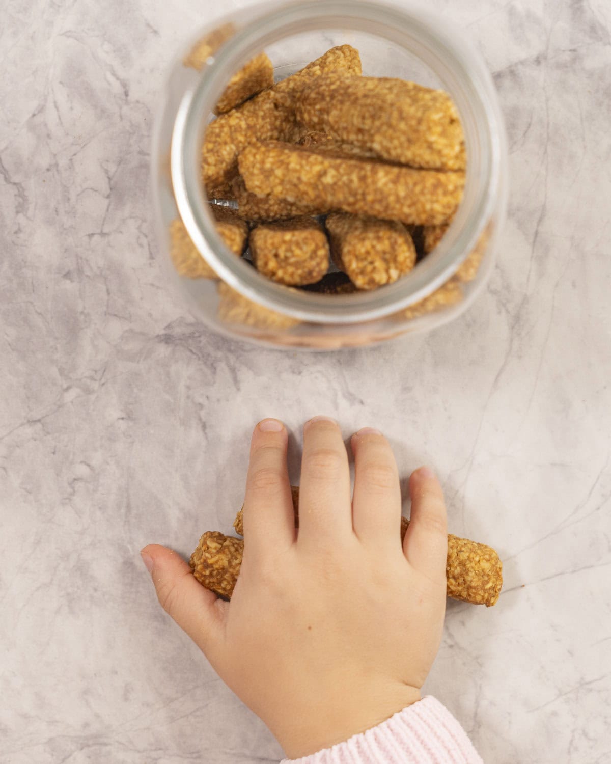 Toddler hand taking a baby rusk from the benchtop next to jar fill of rusks.
