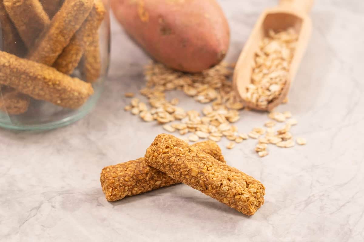 Two baby rusks stacked on each other, wooden spoon with oats, sweet potato and jar with remainder rusks in background.