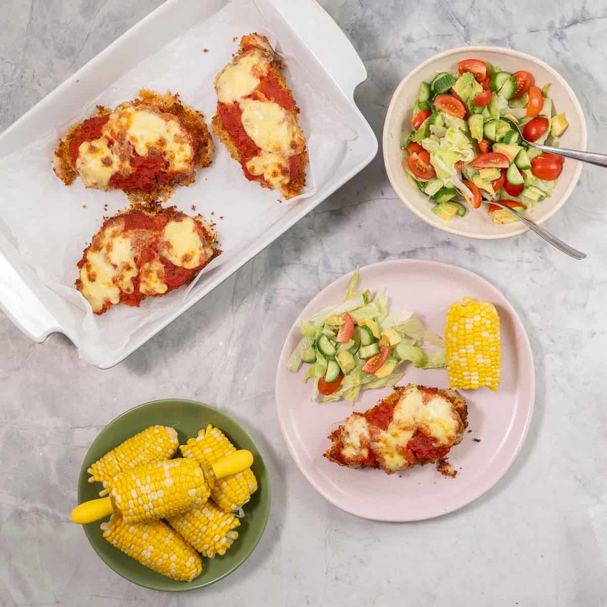 Plate with chicken parmesan breast, fresh salad and corn cob. Bowl with fresh salad, corn on the cob in a bowl and three chicken parmesan in a baking dish.