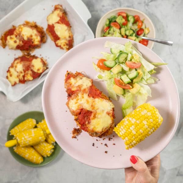 Hand holding a plate with chicken parmesan breast, fresh salad and corn cob.