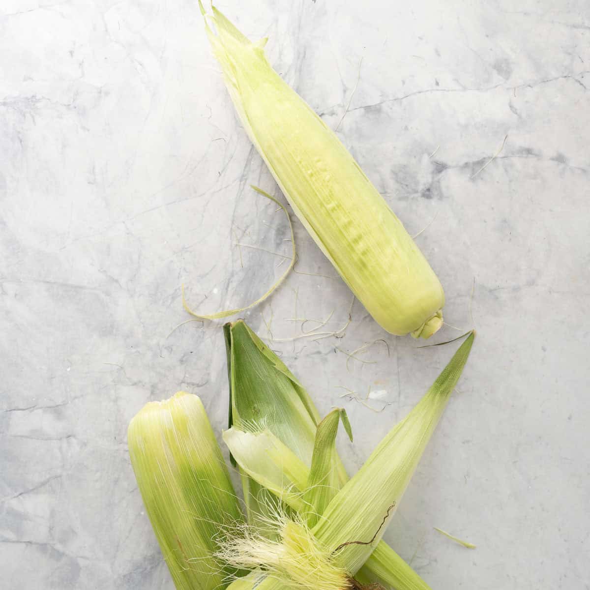 Ear of corn with most of the layers of husk removed on a bench top.