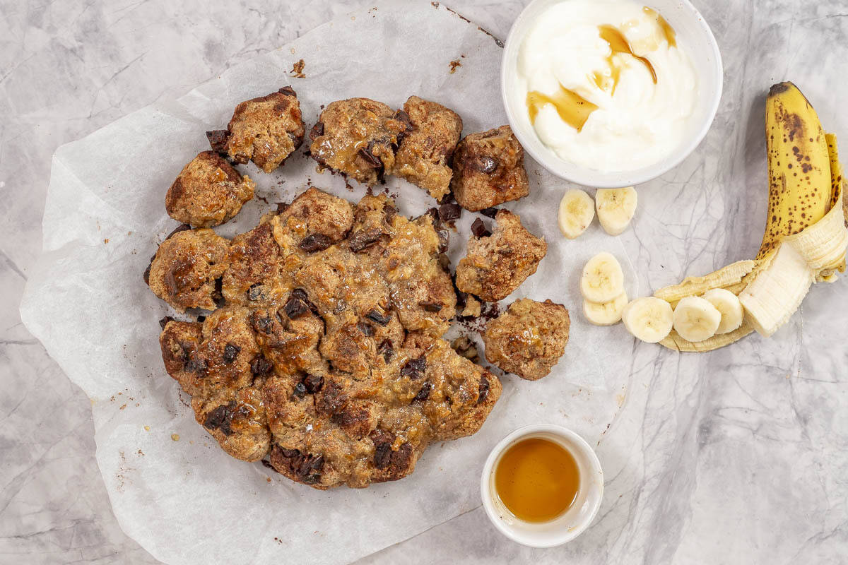 Cooked monkey bread on a piece of baking paper on bench top, desert bowl with yogurt and chopped banana served with monkey bread.