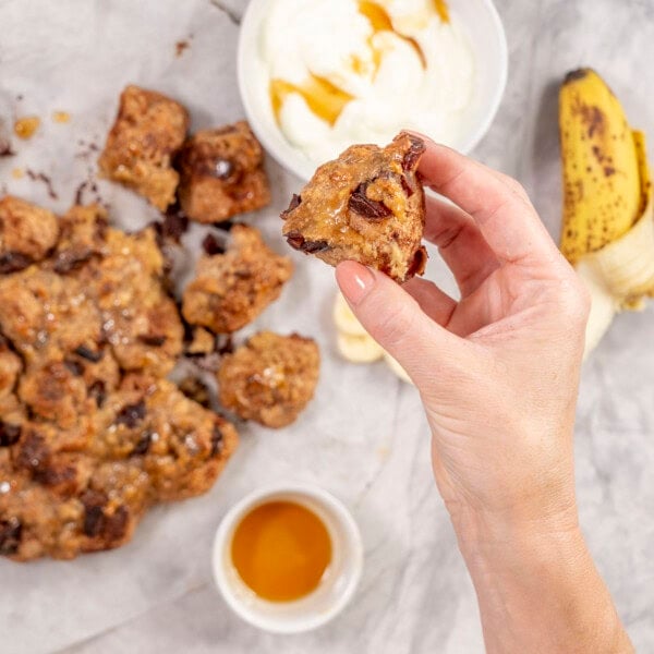 Womans hand holding a piece of monkey bread up to camera with monkey bread, yogurt and banana in background on bench top.