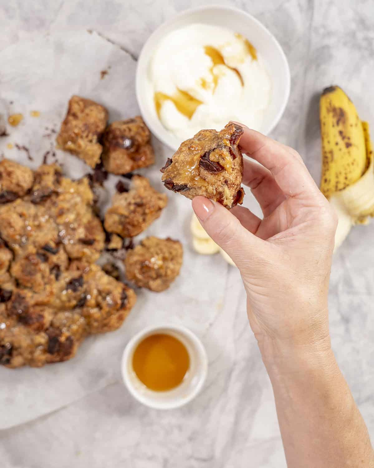 Womans hand holding a piece of monkey bread up to camera with monkey bread, yogurt and banana in background on bench top.