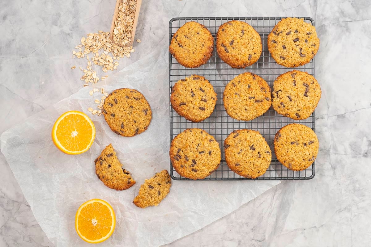 Cooling tray with nine cookies, orange cut in half and cookie broken in half laid on piece of baking paper on benchtop.