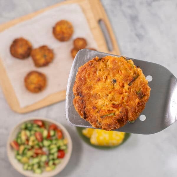 Metal spatula with salmon pattie held up, wooden chopping board with remainder of patties, bowl of fresh salad and corn below.