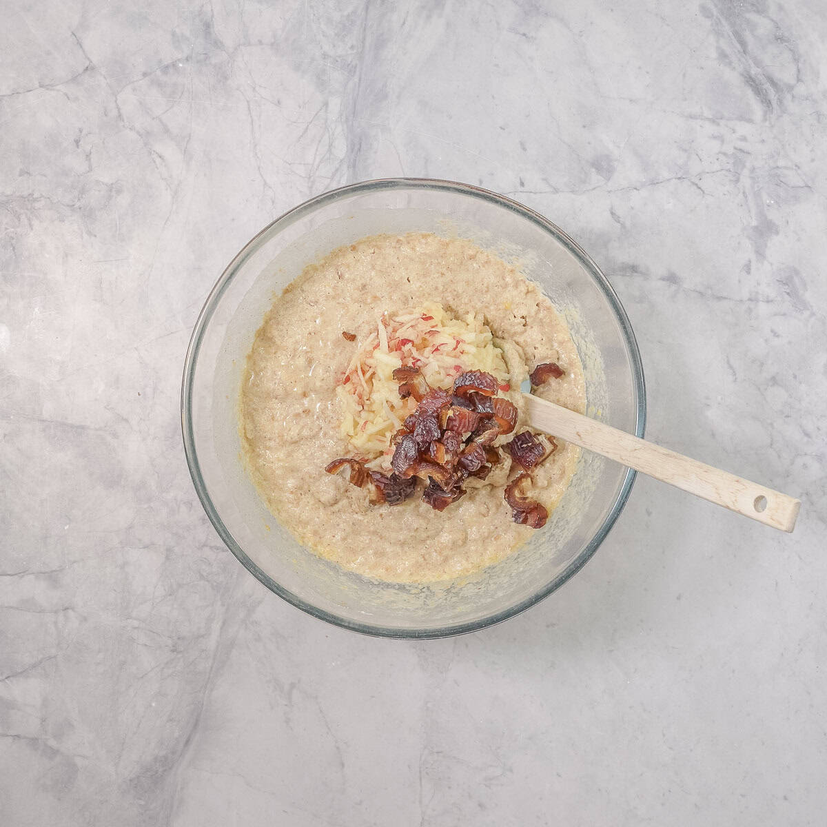 Glass bowl with spatula and mixture for weetbix muffins, grated apple and chopped dates on top.