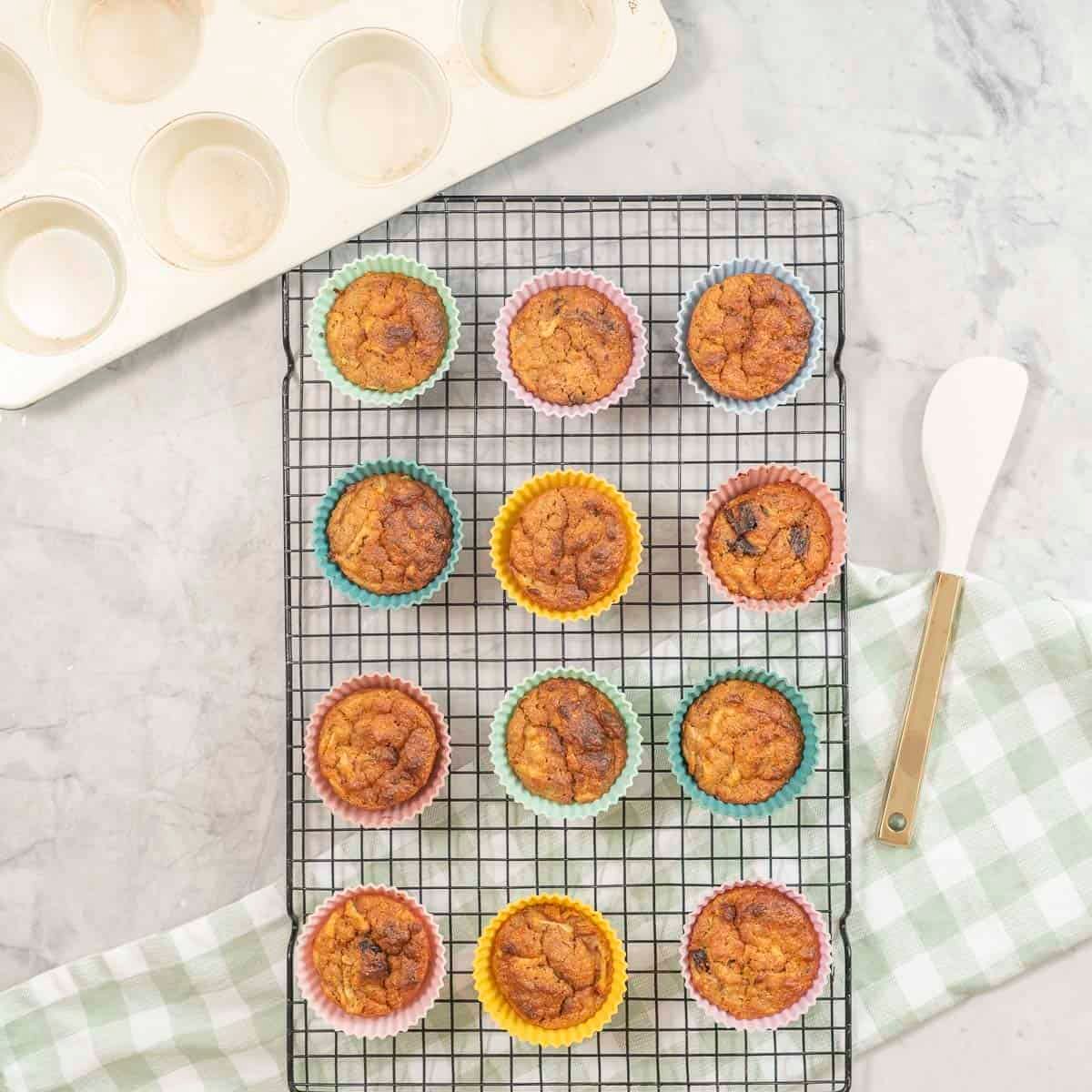 Cooling tray with weetbix muffins on benchtop with checkered tea towel and spatula.