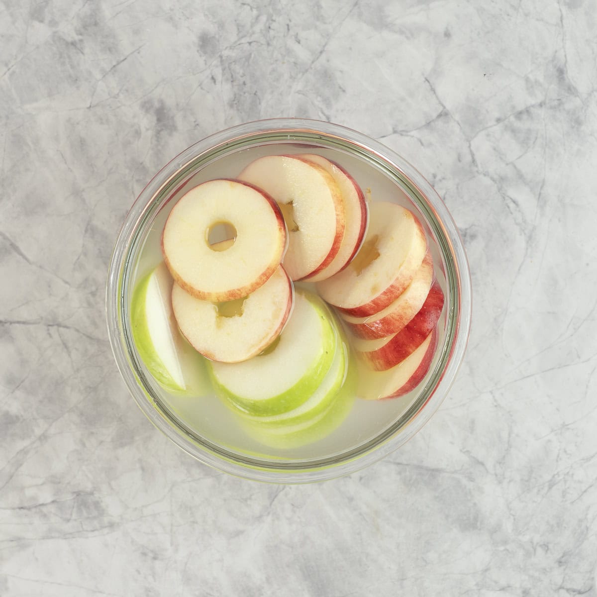 A bowl of red and green apples with core removed sliced like donuts sitting in water.